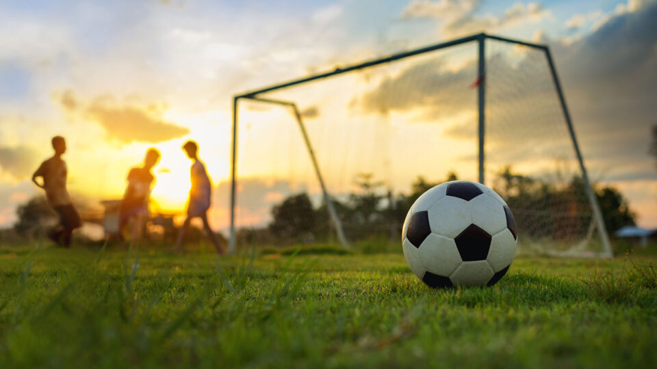 A football in a field next to a goal post at sunset with kids playing in the background