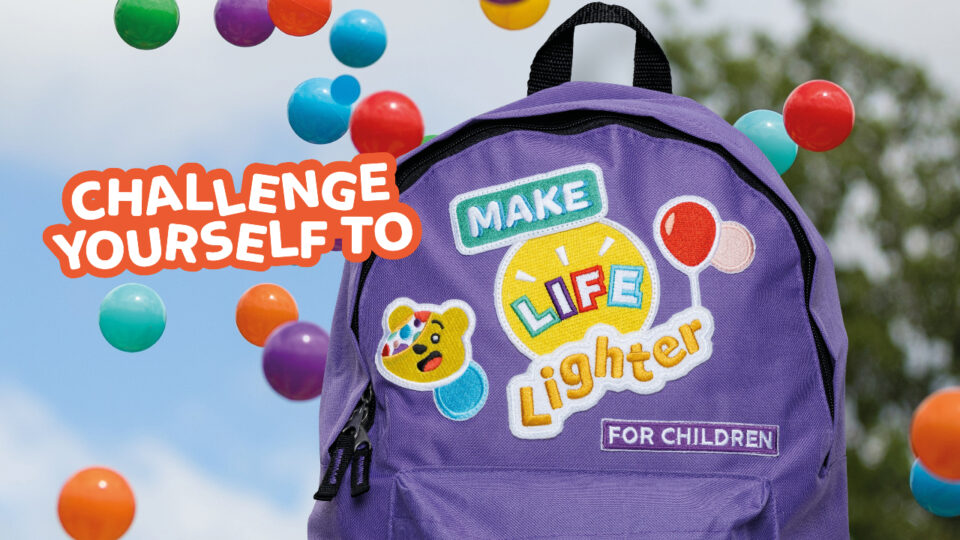 A backpack in front of spotty balls with the message 'challenge yourself to make life lighter for children' embroidered on the backpack