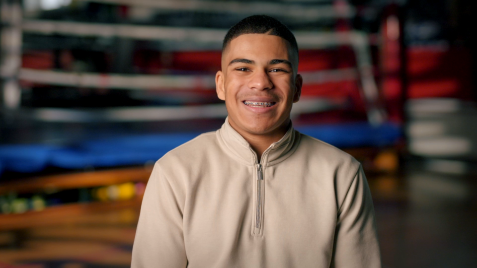 a teenage boy smiling with a boxing ring in the background